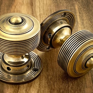 May include: Three antique brass door knobs with a circular design. The knobs feature concentric rings and a small, rounded centre. The brass has a warm, golden hue, set against a wooden surface.