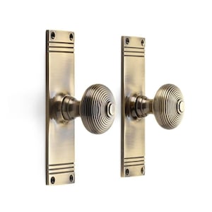 May include: A pair of antique brass door knobs with rectangular backplates. Each knob features a circular, ridged pattern. The backplates have horizontal lines and screw holes. The hardware is set against a white background, ideal for interior design.