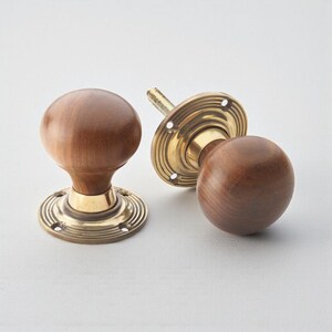 May include: Two wooden door knobs with brass bases. The knobs are a warm brown colour and have a rounded shape. The brass bases have a circular design with multiple grooves. One knob is detached, showing the screw.