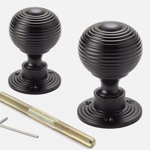 May include: Two black door knobs with a ribbed, spherical design. The knobs sit on a round base and are accompanied by a threaded rod and an Allen key. Hardware for home decor.