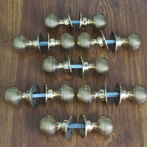 May include: A selection of antique brass door knobs with a ribbed design, arranged on a brown wooden surface. Each knob features a round shape, a central spindle, and two backplates. The knobs are made of brass.