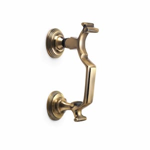 May include: A brass door knocker with a curved handle and circular backplates. The knocker has an antique finish and is mounted on a white surface. The design is ornate and traditional, suitable for exterior doors.