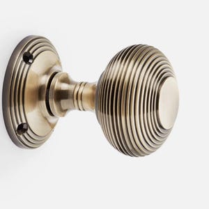 May include: A brass door knob with a circular backplate and an oval handle. The handle has a grooved design, and the backplate has a simple style. The hardware is mounted on a white surface. A classic piece of door hardware.