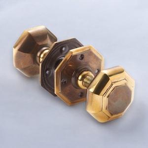 May include: Three antique brass door knobs with an octagonal design. The knobs have a polished finish, while the backplates show an aged patina. The door hardware is displayed against a light grey background. The knobs are likely for a door.