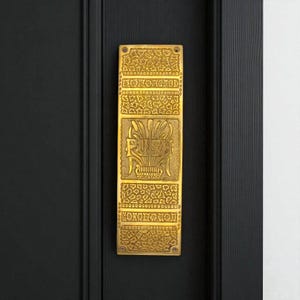 May include: A rectangular brass push plate with intricate floral and geometric designs. The word "PUSH" is centrally displayed in a decorative font. The plate is fixed to a black wooden door.