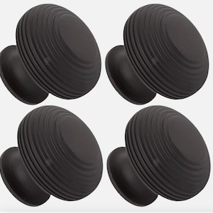 May include: Four black cabinet knobs with a ridged, circular design. Each knob has a rounded face and a smaller, circular base. The knobs are made of a dark, matte material and are evenly spaced against a light background.