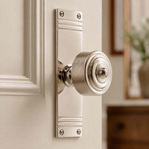 May include: A polished nickel door knob set featuring a round, ribbed handle attached to a rectangular backplate. The backplate has horizontal grooved detailing at the top and bottom and is secured to a white door with four visible metal screws.