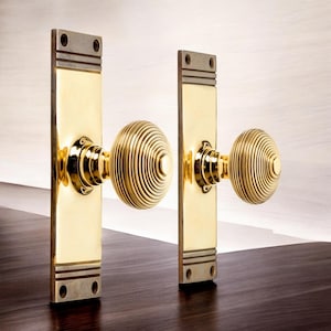 May include: A pair of polished brass door handles. Each handle has a rectangular backplate with a decorative top and bottom, and a round, ridged knob. The handles are set against a light background and a dark wooden surface.