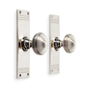 May include: Two polished nickel door knobs with matching rectangular backplates. The knobs have a circular, grooved design. The backplates have horizontal lines and screw holes. The hardware is designed for interior doors.