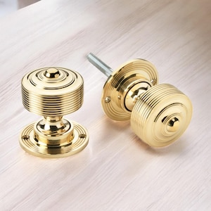 May include: Two polished brass door knobs. The knobs have a circular base and a cylindrical handle with horizontal grooves. One knob is disassembled, showing the threaded rod. The hardware is set against a light wood grain surface.