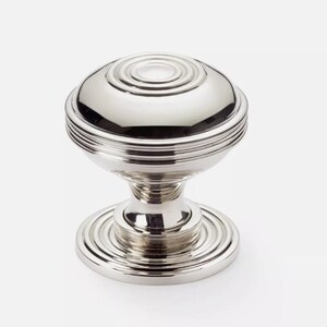 May include: A shiny silver cabinet knob with a circular, layered design. The knob has concentric circles on the top and base, and horizontal ridges around the middle. The hardware is designed for furniture and cabinetry.