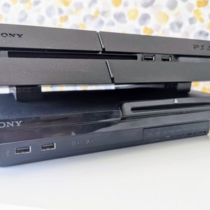 May include: Two black Sony PlayStation consoles stacked on top of each other. The top console is a PlayStation 4 and the bottom console is a PlayStation 3.