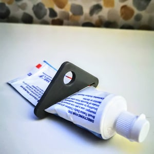 May include: A black plastic toothpaste tube squeezer with a hole in the centre. The squeezer is holding a blue and white toothpaste tube with the text "DIRECTIONS FOR USE: Brush Twice Daily Children of 6 years younger, use a pea size amount for dentist or doctor. intake of fluoride from other brushing to minimise swallowing PL-50-100 Swi GUILDFORD GAZ B COLGATE-PALMOLIVE Call bee: 00-800-32 SAVE MATER".