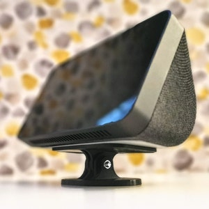 May include: A black smart speaker with a grey fabric speaker grille and a white bezel. The speaker is mounted on a black stand with a circular base.