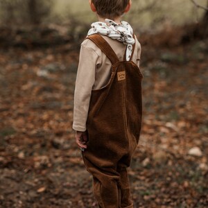 Op de afbeelding: Een jong kind draagt een bruine corduroy broek met bretels en een witte en bruine gestreepte sjaal. Het kind staat op een boomstronk in een bosrijk gebied.