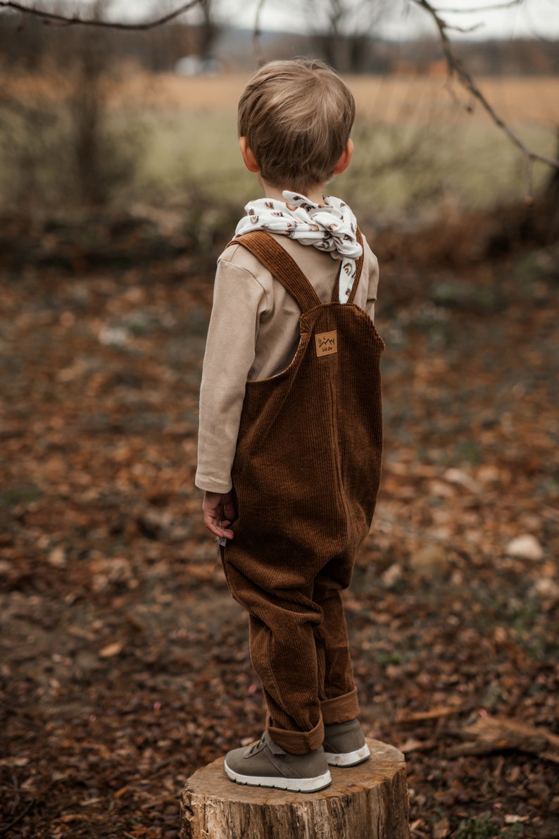 Op de afbeelding: Een jong kind draagt een bruine corduroy broek met bretels en een witte en bruine gestreepte sjaal. Het kind staat op een boomstronk in een bosrijk gebied.