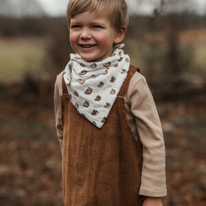 Op de afbeelding: Een jong kind draagt een bruine corduroy overall en een witte bandana met een regenboogpatroon. Het kind lacht en kijkt naar rechts.