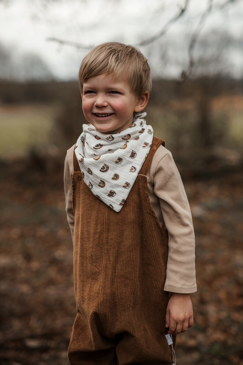Op de afbeelding: Een jong kind draagt een bruine corduroy overall en een witte bandana met een regenboogpatroon. Het kind lacht en kijkt naar rechts.