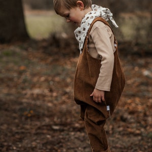Op de afbeelding: Een jong kind draagt een bruine corduroy overall met een witte bandana met een bruin printje. Het kind staat op een boomstronk in een bosrijk gebied.