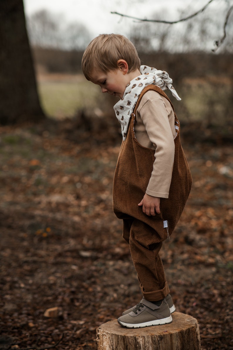 Op de afbeelding: Een jong kind draagt een bruine corduroy overall met een witte bandana met een bruin printje. Het kind staat op een boomstronk in een bosrijk gebied.
