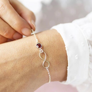 May include: A silver infinity bracelet with a small, dark red gemstone. The bracelet is on a person's wrist.