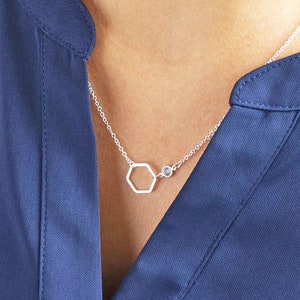 May include: A silver necklace with a hexagonal pendant and a small round gemstone. The necklace is on a model wearing a blue shirt.