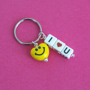 Pode incluir: Um chaveiro prateado com um charme de coração amarelo com cara sorridente e três contas de letras brancas que soletram "Eu te amo". A conta de coração no meio tem um coração vermelho.