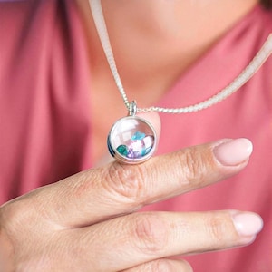 Può includere: Una collana con pendente in argento con un ciondolo rotondo e trasparente pieno di pietre preziose colorate. Il ciondolo ha una fascia blu. La collana è esposta su un dito su sfondo rosa.