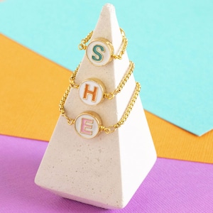 Può includere: Tre braccialetti a catena dorati con ciondoli a lettera smaltati. Le lettere compongono la parola "SHE" su uno sfondo bianco con lettere colorate. Le lettere sono verde, arancione e rosa.