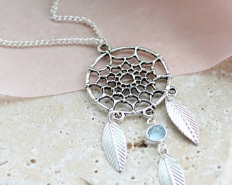 May include: Silver dreamcatcher necklace with a blue gemstone and three feathers. The necklace is on a light brown surface.