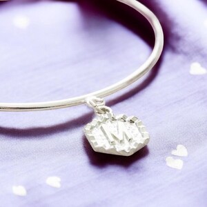 May include: A silver bangle bracelet with a charm that has the letter "M" embossed on it. The charm is a hexagonal shape with a textured surface.