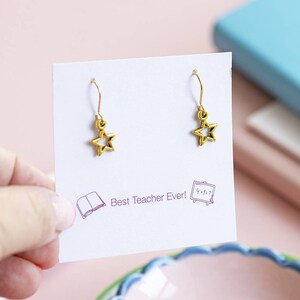 May include: A pair of gold star earrings on a white card with the text "Best Teacher Ever!" and illustrations of a book and a chalkboard.