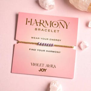 Könnte beinhalten: Ein goldfarbenes Perlenarmband mit violetten Perlen, präsentiert auf einer rosa Karte. Auf der Karte steht "HARMONY BRACELET", "WEAR YOUR ENERGY" und "FIND YOUR HARMONY". Auf der Unterseite der Karte steht "VIOLET AURA JOY".