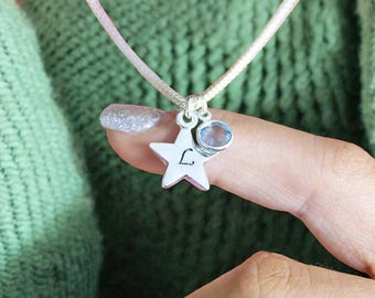 May include: A silver star charm necklace with a light blue gemstone and the letter "L" engraved on the star. The necklace is on a light pink cord.