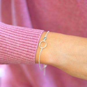 May include: A silver bracelet with a small open circle and a light blue gemstone. The bracelet is attached to a fine chain. The bracelet is shown on a wrist, against a pink background.