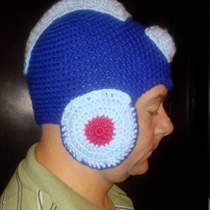 May include: A blue and white crocheted hat with a red circle in the center of the ear flaps. The hat is worn by a person with a green striped shirt.