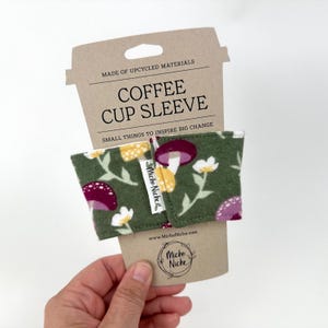 May include: A green coffee cup sleeve with a mushroom and floral pattern. The sleeve is made of upcycled materials and has the text "COFFEE CUP SLEEVE" printed on the attached cardboard. The brand name "Miche Niche" is also visible.