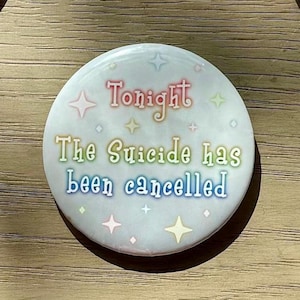 May include: A white round button with rainbow-coloured text that reads "Tonight The Suicide Has Been Cancelled" and a white background with small rainbow-coloured stars.