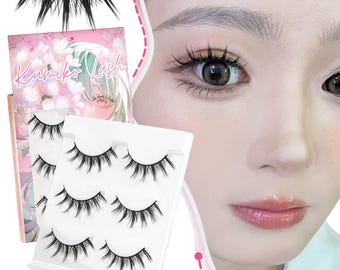 Könnte beinhalten: Das Bild zeigt mehrere Sets falscher Wimpern, zusammen mit einer rosa-weißen Schachtel mit der Aufschrift "Kumiko Lash". Es gibt auch eine Nahaufnahme eines Auges mit angebrachten falschen Wimpern.