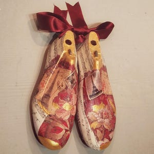 Puede incluir: Un par de formas de zapatos de madera decoradas con papel vintage, incluyendo notas musicales, sellos y patrones florales. Las formas están atadas juntas con una cinta de satén burdeos.