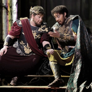 May include: Two men wearing elaborate costumes, one with a crown, are seated on a stage. They are both wearing gold armor and robes. The man on the left is wearing a red robe with gold trim, while the man on the right is wearing a green robe with gold trim. They are both looking at each other and talking.