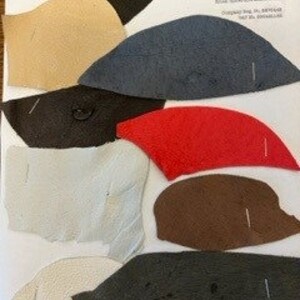 May include: A selection of fabric swatches in diverse colours such as black, beige, navy, red, brown, and white. Each swatch has a unique shape and size, with pins attached. The swatches are arranged on a white background with text at the top.