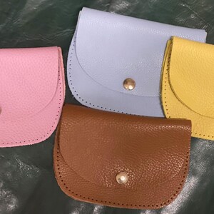 Könnte beinhalten: Vier kleine Leder-Münzbörsen in Pastellfarben: Rosa, Hellblau, Gelb und Braun. Jede Börse hat eine gewölbte Klappe, die mit einem goldfarbenen Druckknopf befestigt ist. Die Börsen sind auf einer dunkelgrünen Oberfläche angeordnet.