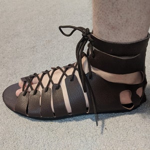May include: Brown leather sandals with a lace-up design and an ankle strap. The sandals have a cut-out design on the back and are worn on a foot.