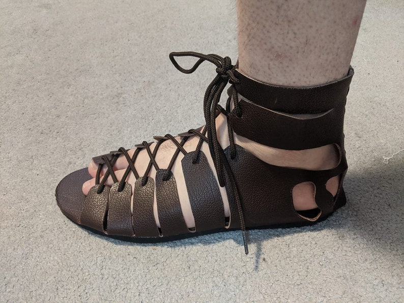 May include: Brown leather sandals with a lace-up design and an ankle strap. The sandals have a cut-out design on the back and are worn on a foot.