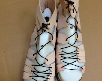 May include: A pair of beige leather sandals with black laces. The sandals have a closed toe and an open heel. The laces are tied in a criss-cross pattern.
