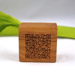 May include: A small, square wooden block with a QR code etched into one side. The block is a warm brown colour and appears to be made of wood. A green leaf is visible in the background.