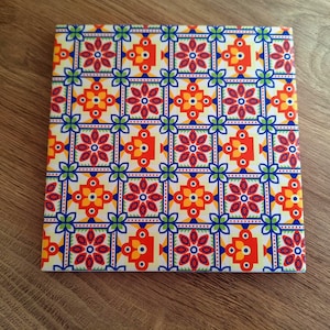 Puede incluir: Azulejo decorativo cuadrado con un patrón repetitivo de formas florales y geométricas rojas, naranjas, amarillas y azules sobre un fondo blanco. El azulejo está sobre una superficie de madera.