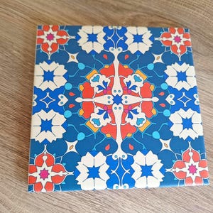 May include: Square decorative tile with a vibrant floral pattern. The design features a blue background with white, orange, and red floral motifs. The tile is likely ceramic and suitable for home decor.