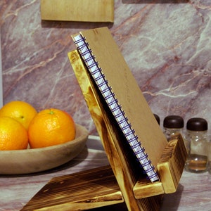 Può includere: Un porta-ricettario in legno con un quaderno marrone con una rilegatura a spirale blu. Il supporto è in legno scuro con una finitura rustica. Il supporto è appoggiato su un piano di lavoro in marmo.